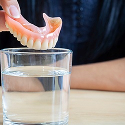 Fingers holding upper set of dentures over a glass of clear fluid on a table